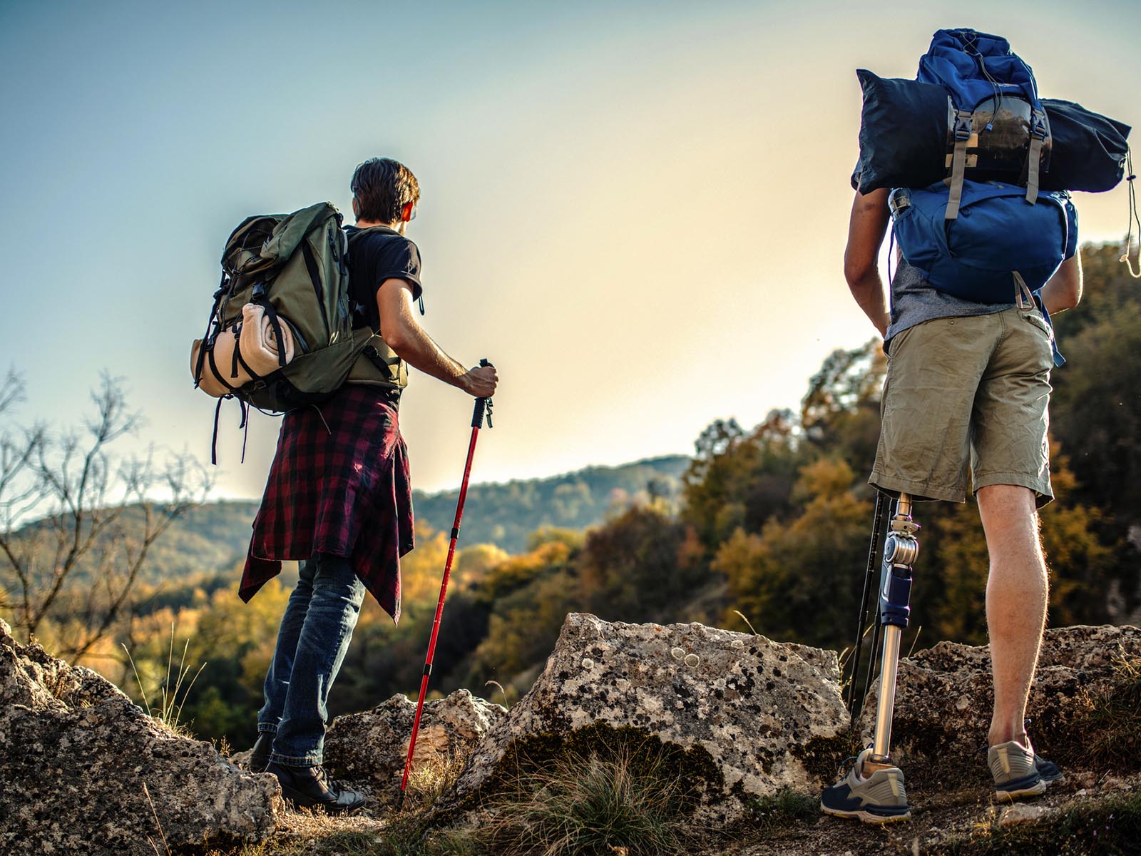 2 people hiking in the mountains with heavy backpacks on their backs. They are looking out into the distance. One of the hikers is amputee